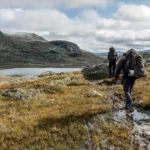 Dois praticantes de trekking caminhando por uma trilha de montanha com mochilas e bastões próximos a um lago.